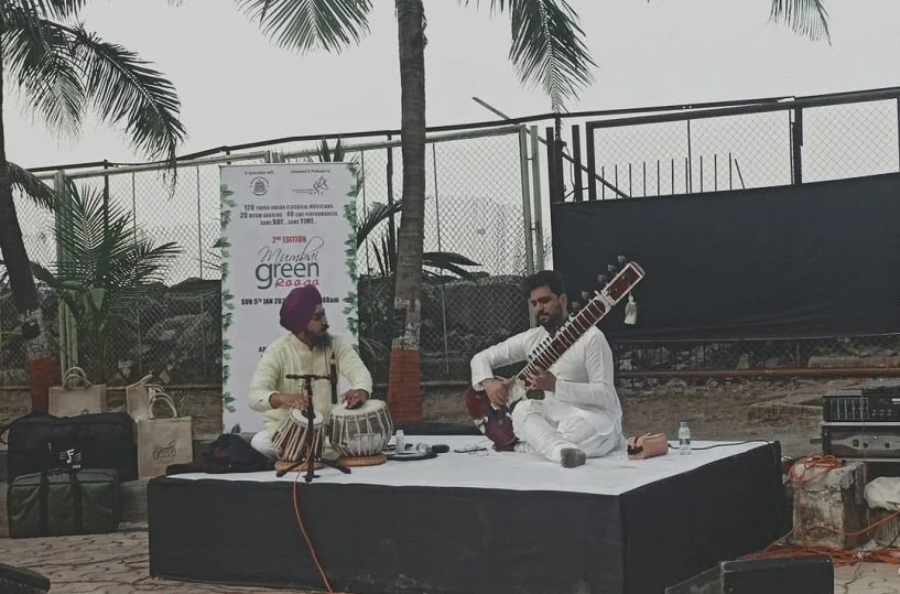 Two musicians performing outdoors on a makeshift stage, one playing tabla and the other playing a sitar, with palm trees and a fence in the background.