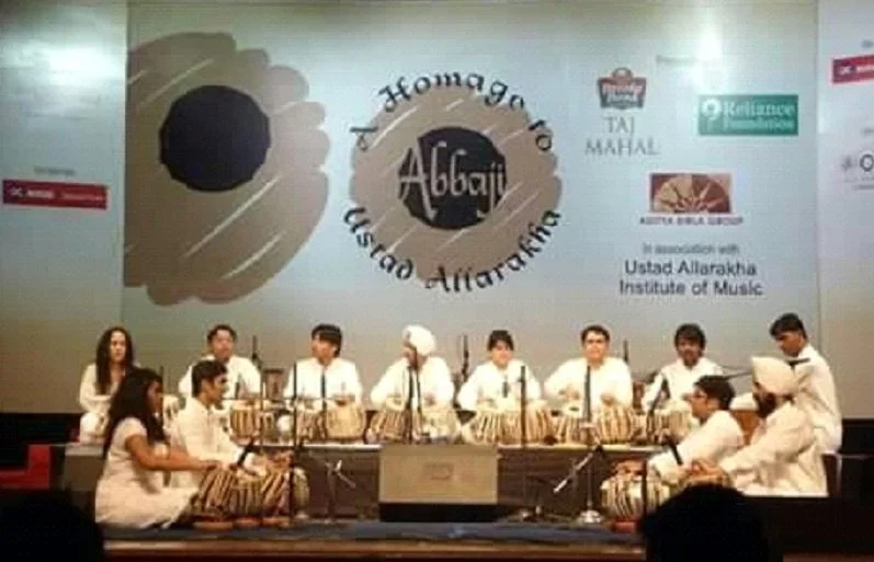 A group of young performers in white traditional Indian attire playing tabla drums on stage at an event, with a large projected backdrop featuring logos and text related to Ustad Allahrakha Institute of Music.