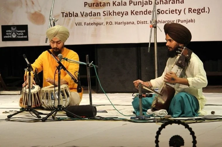 Two musicians dressed in traditional Indian attire performing on stage; one playing the tabla and the other playing a violin, with microphones set up in front of them.