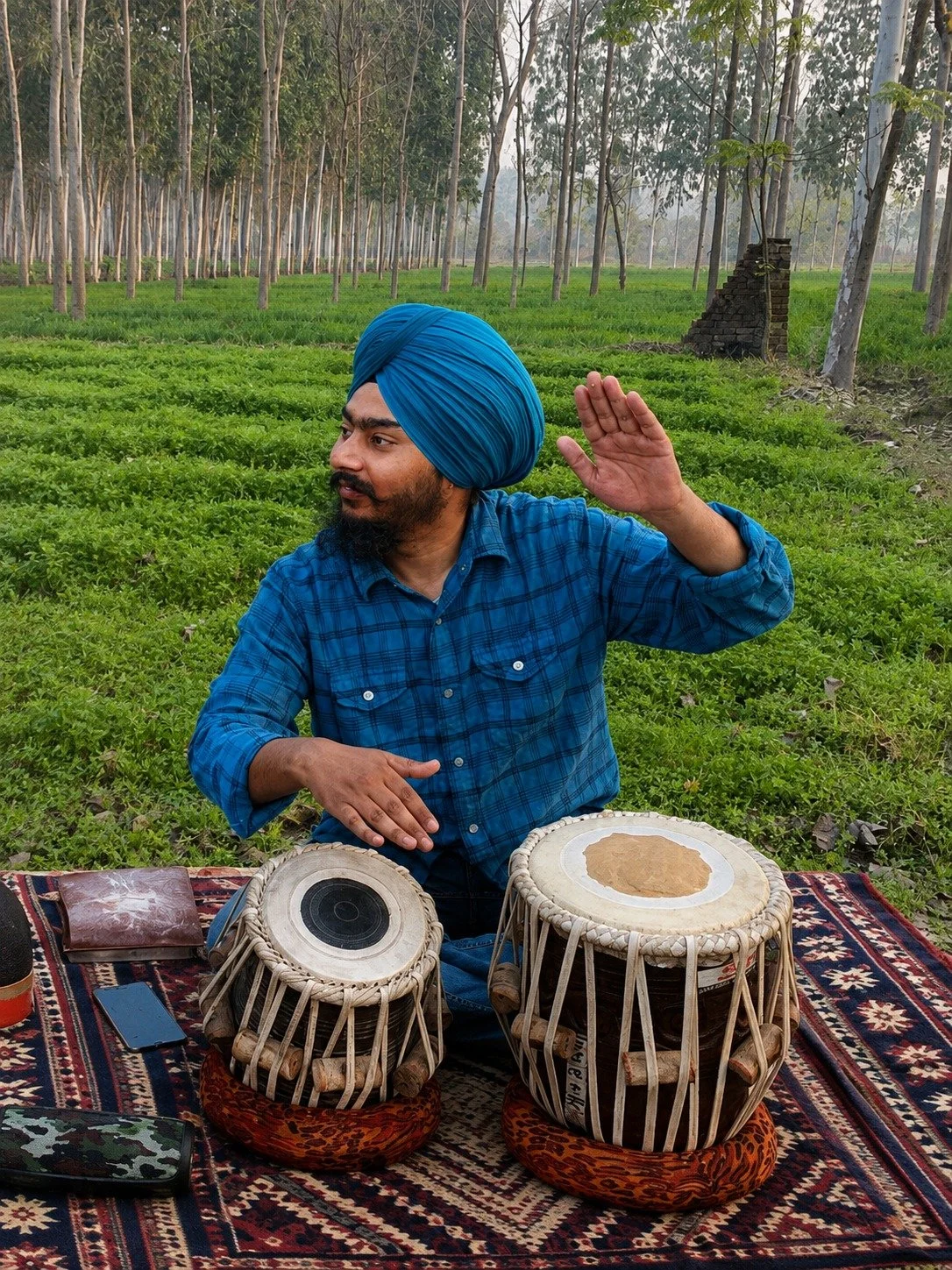 A man wearing a blue turban and checkered shirt sitting on a patterned rug outdoors, playing traditional drums with a lush green forest in the background.