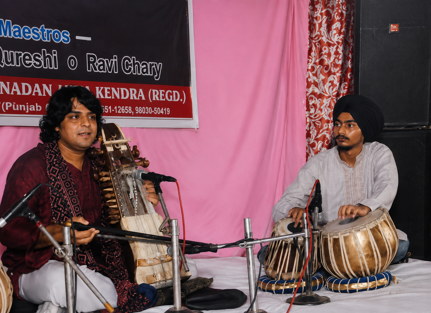 Two men are seated on a stage, one playing a sitar and the other playing tabla drums. They are performing at an event with a pink backdrop and a banner behind them.