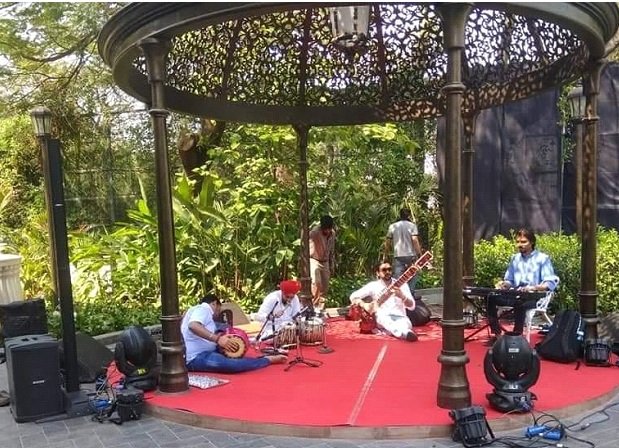 A musical performance on a small outdoor stage with traditional decorations, featuring four musicians playing instruments, surrounded by greenery and audience.
