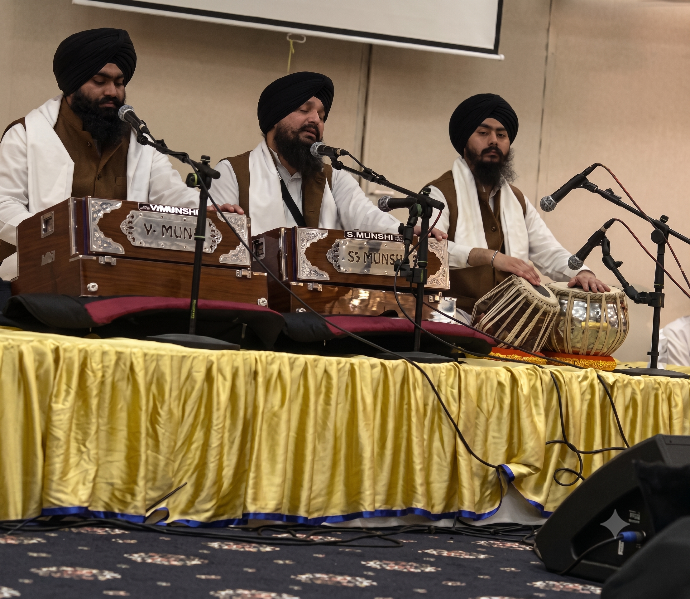 Three men wearing turbans and traditional attire are seated behind a decorated wooden tabla, performing at a cultural event on stage. Microphones are positioned in front of them, and a yellow curtain drapes the front of the stage.