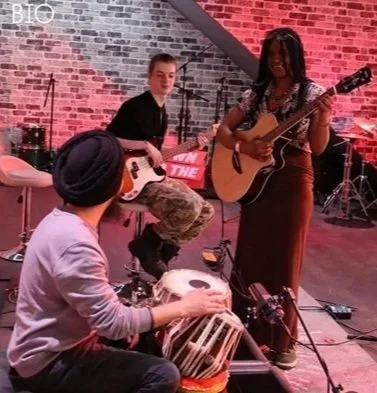 Three musicians performing on stage: a woman with a guitar singing, a man playing an acoustic guitar, and a person playing a drum set in front of a brick wall.