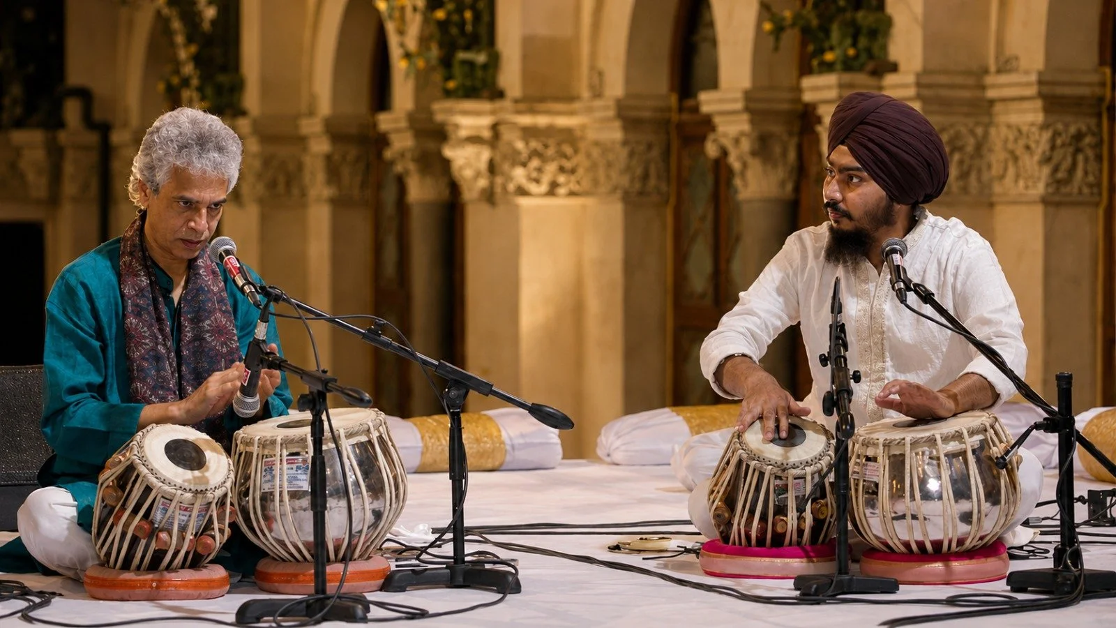 Two men sit on the floor playing tabla drums with microphones in front of them, in a decorated ornate hall.