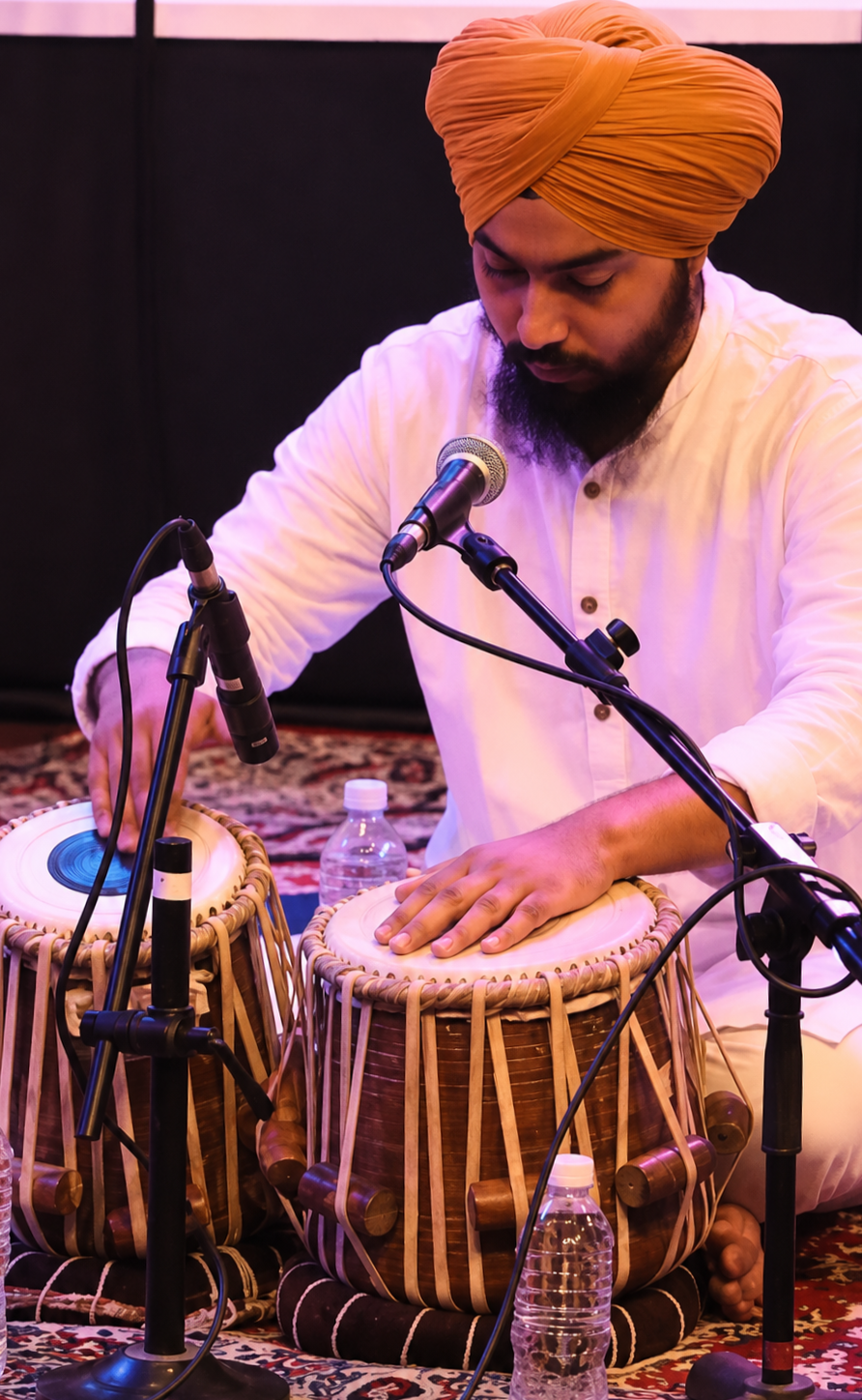 A man wearing a tan turban and white shirt is sitting on a patterned rug, playing two traditional drums with his hands. There is a microphone positioned in front of him, and a water bottle is on the rug nearby.