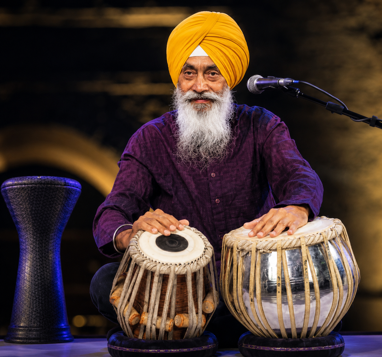 An elderly man with a white beard and yellow turban playing two traditional Indian drums, sitting in front of a microphone, with a dark, blurred background.