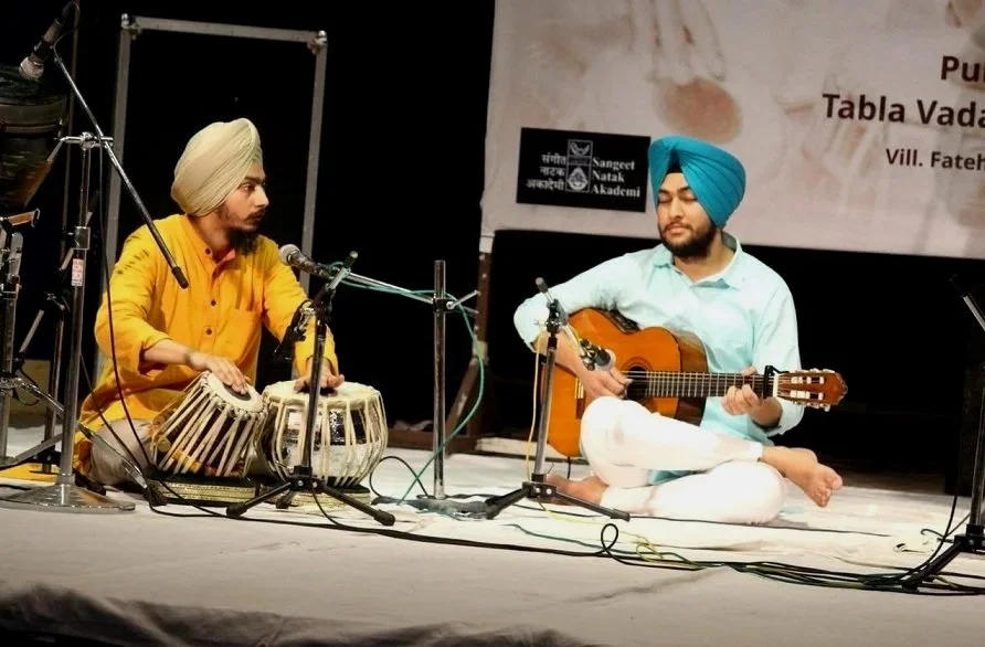 Two men performing on stage; one is playing a tabla and the other is playing a guitar, both wearing traditional Punjabi turbans and seated cross-legged.