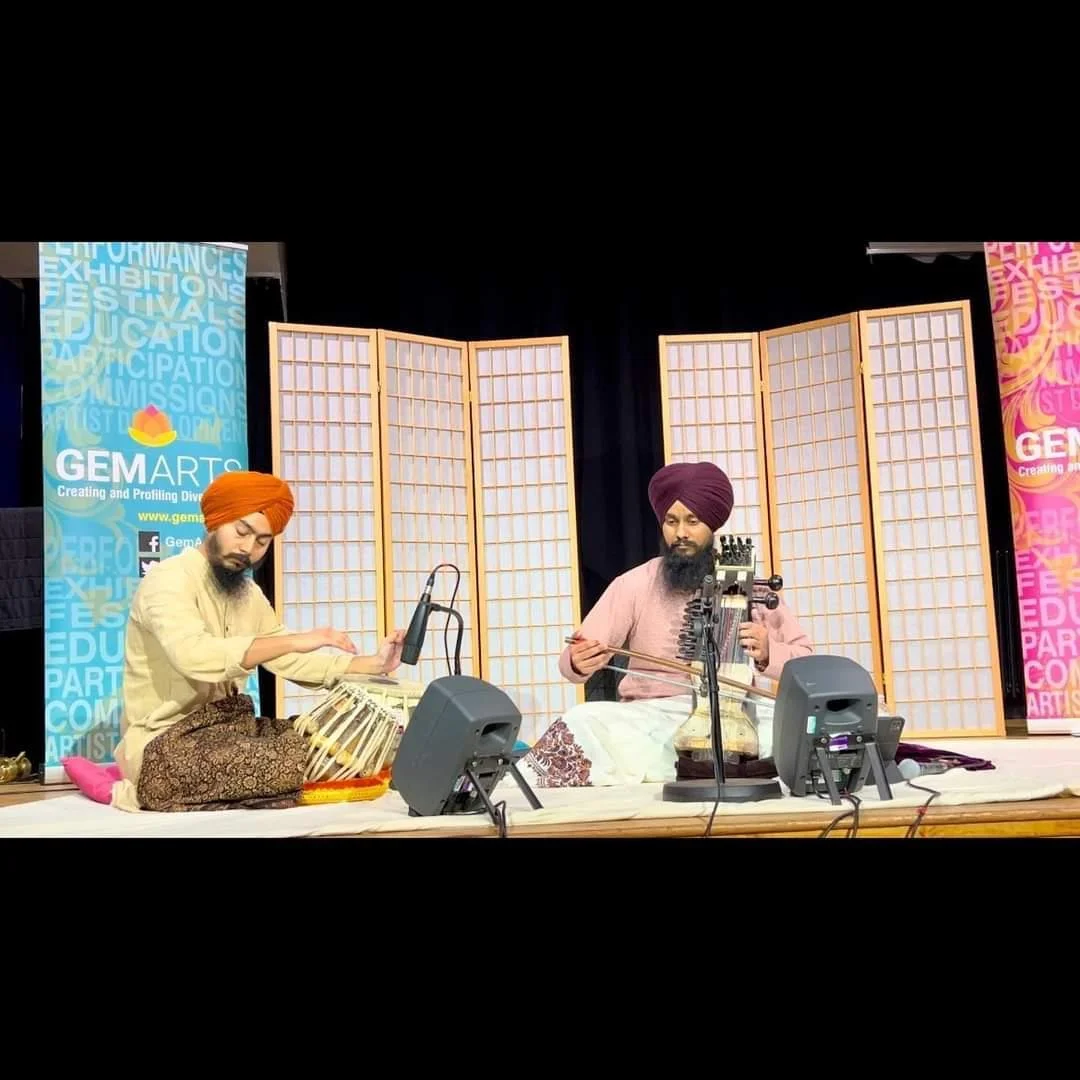 Two men in traditional Sikh attire playing classical Indian instruments on stage with folding screens and GEMARTS banners in the background.