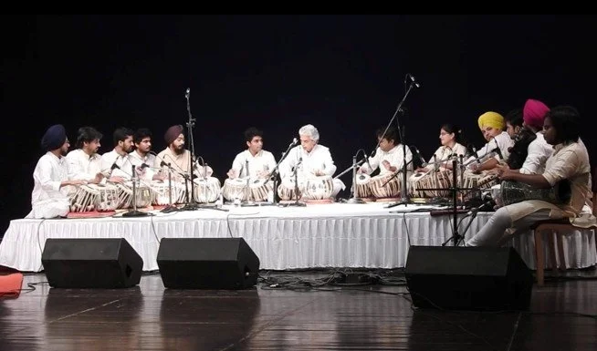 Group of Indian musicians performing on stage, playing traditional instruments under stage lighting.