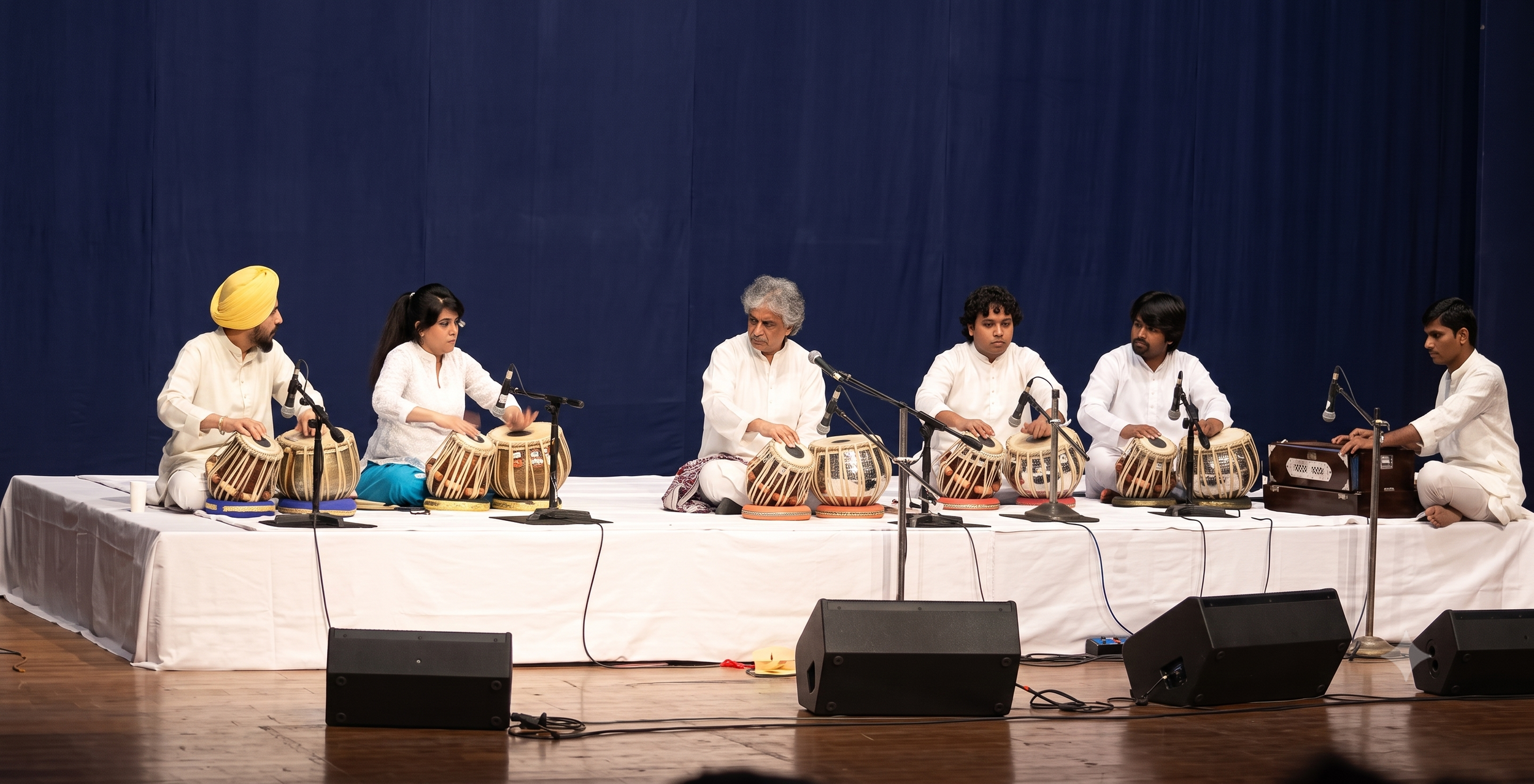 Group of six people sitting on stage playing traditional Indian drums called tabla, with microphones set up in front of them, against a dark blue curtain background.
