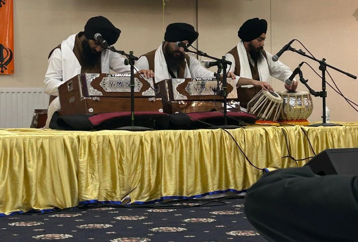 Three men wearing turbans and traditional attire playing musical instruments on a stage with a yellow tablecloth and microphones.