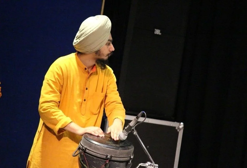 A man wearing a yellow kurta and a turban playing a drum with a microphone attached, on stage with a dark background.