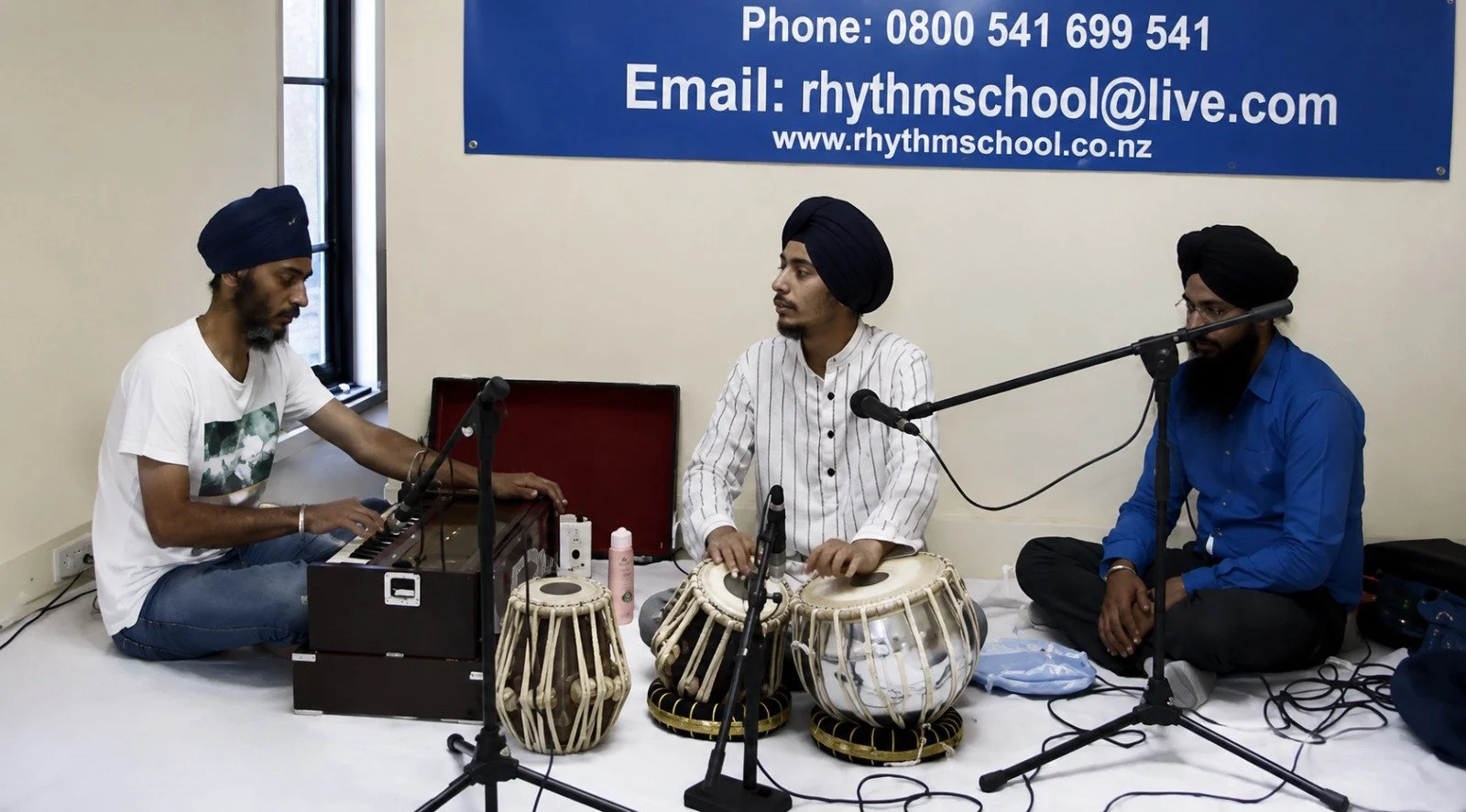 Three men wearing turbans, sitting on the floor, playing musical instruments. The man on the left is playing a harmonium, the man in the middle is playing tablas, and the man on the right appears to be singing or listening. There is a blue sign with contact information on the wall behind them.