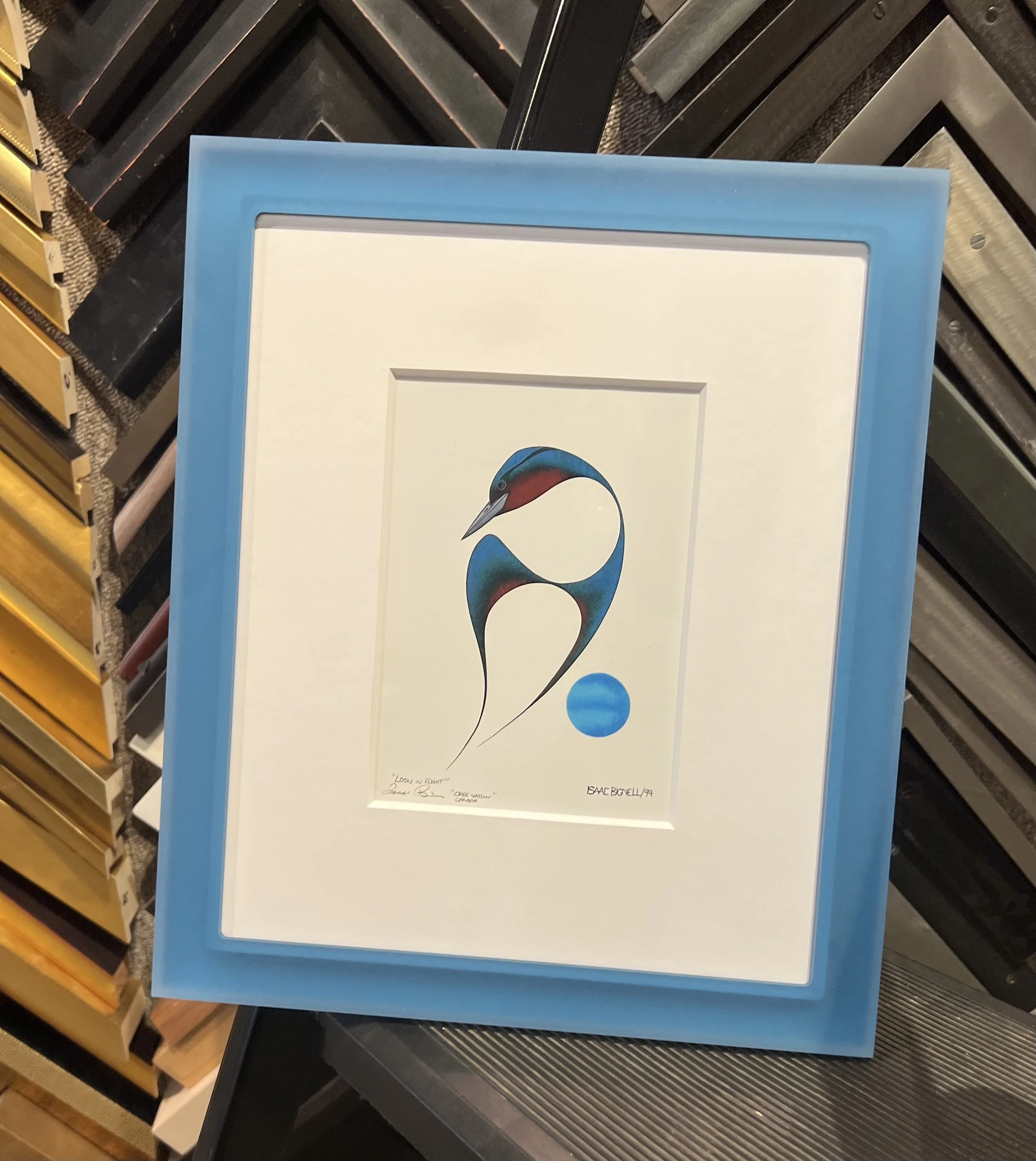Framed artwork featuring an abstract painting of a bird with a red beak and blue body, placed on a table in a store.