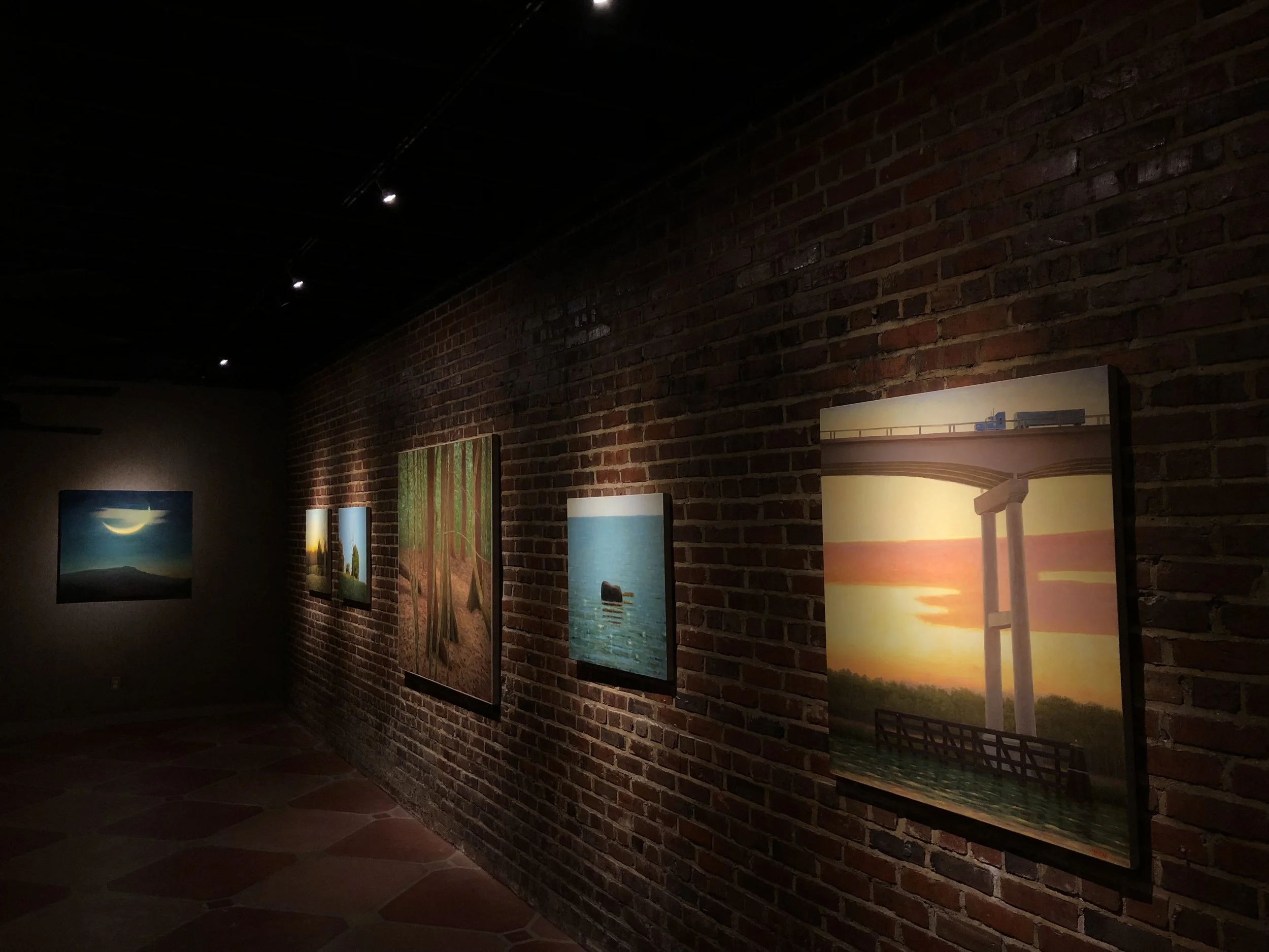 Art gallery with paintings of nature scenes hanging on a brick wall, dimly lit with spotlights.