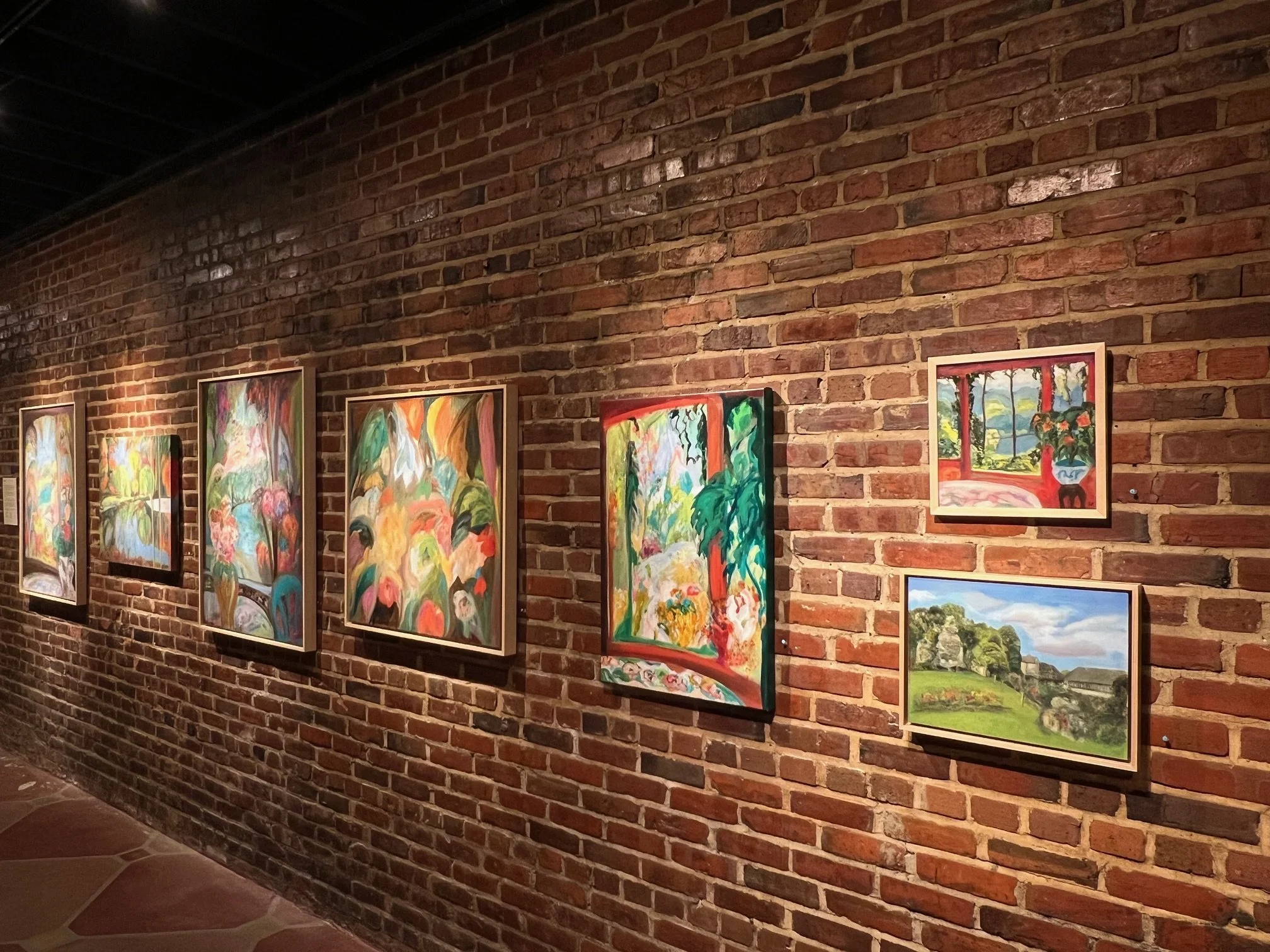 Artwork displayed on a brick wall in an art gallery, featuring colorful, abstract paintings of natural scenes such as trees and landscapes.