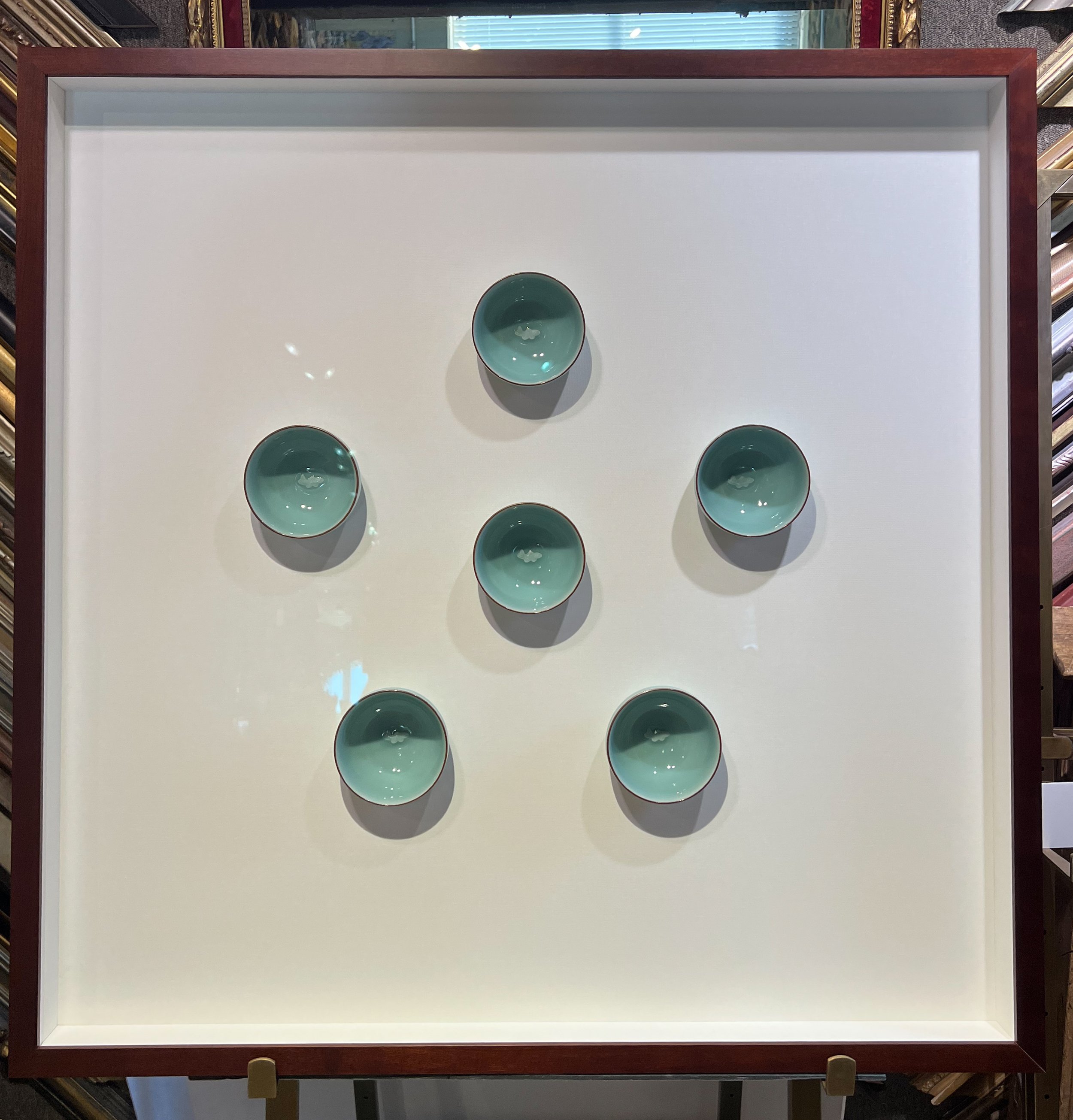 From a top-down view, a display case with a dark wooden frame houses five turquoise bowls with a glossy glaze, arranged in a staggered pattern on a white surface inside the case.