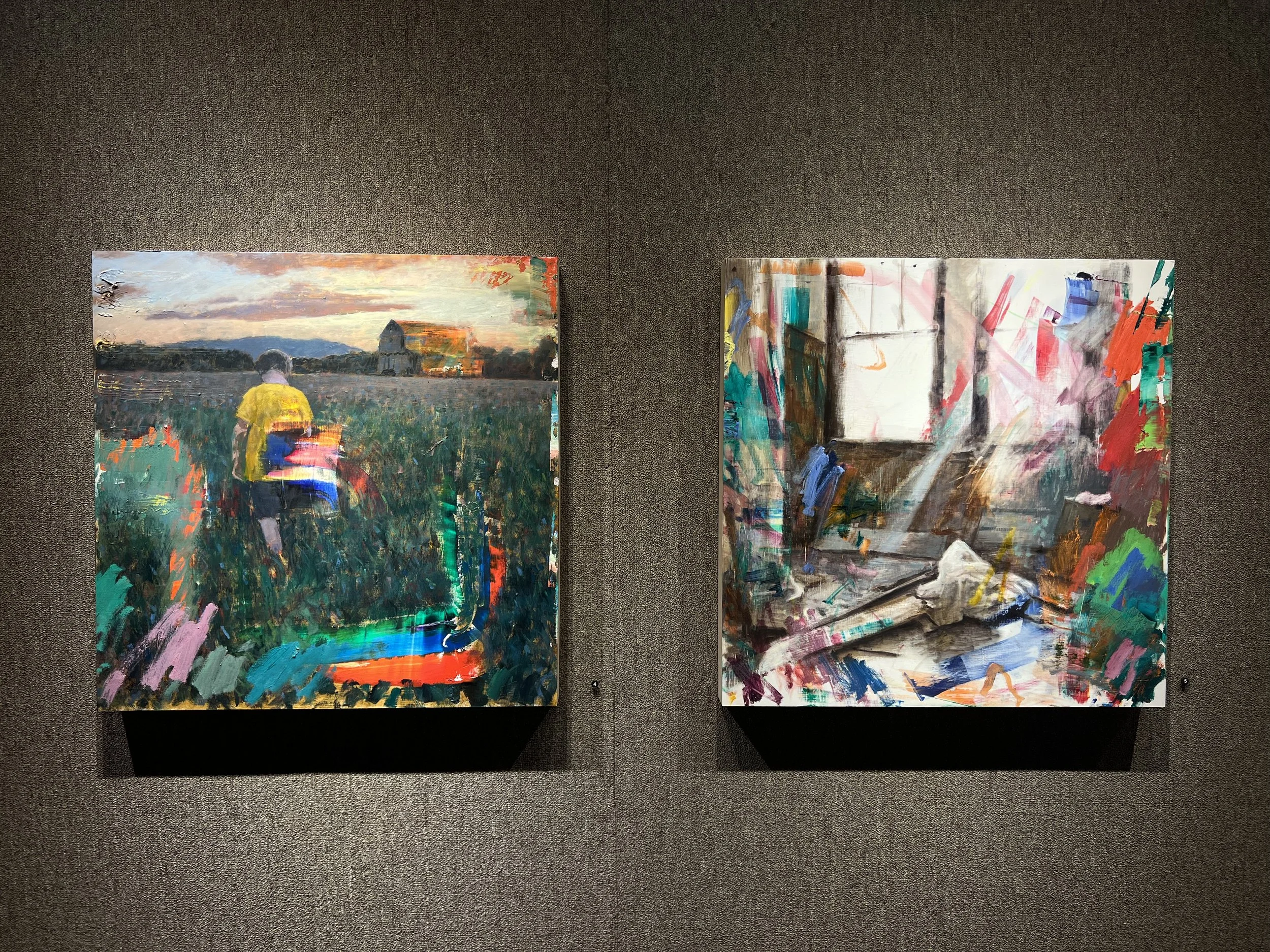 Two abstract paintings displayed side by side on a dark carpeted floor. The left painting depicts a landscape with a person in a yellow shirt in a field during sunset, with soft pastel skies and distant mountains. The right painting features colorful, expressive brushstrokes and abstract shapes.