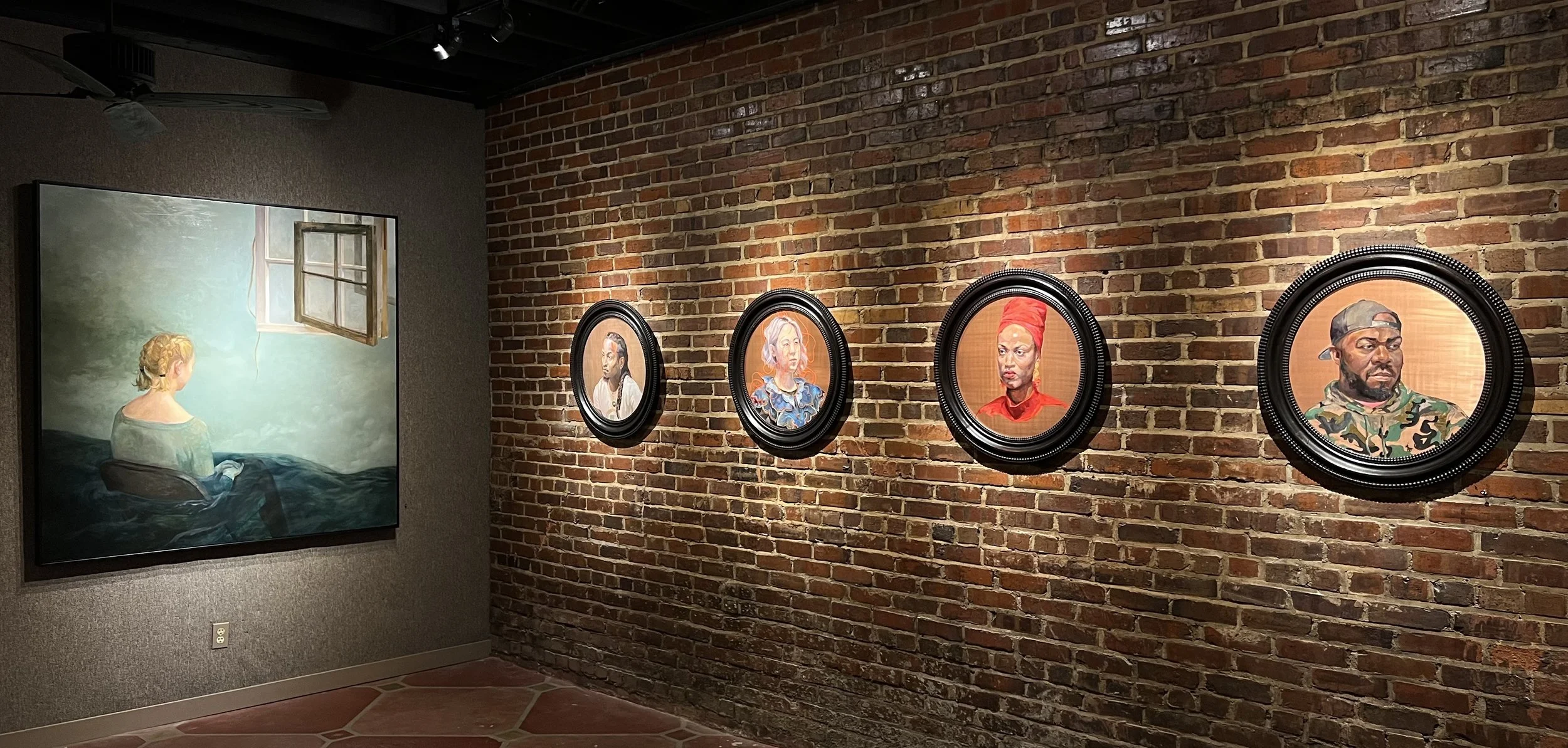 Art gallery wall with five framed portraits of diverse people hung on a brick wall, and a large painting of a woman looking out a window on a gray wall.