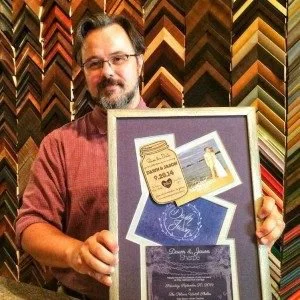 A man with glasses and a beard, wearing a red shirt, holding a framed display that includes a photograph of a wedding couple and various decorative items, in a background of framed picture mats.