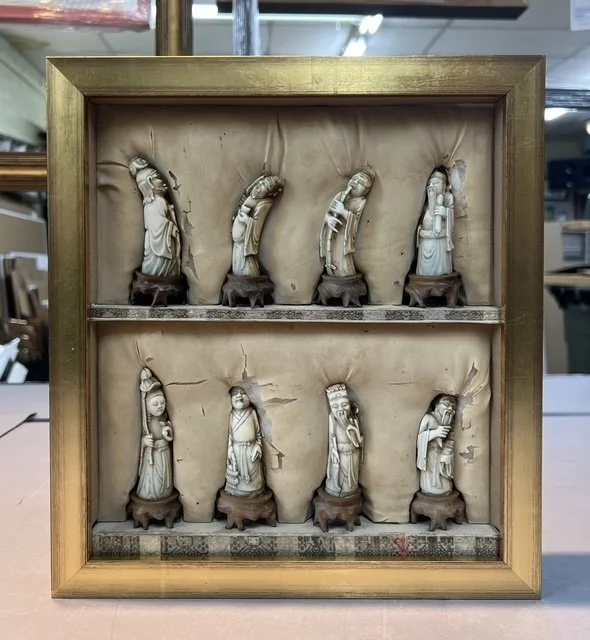 A framed display of eight small carved ivory figurines, each depicting a different religious figure or saint, arranged in two rows on wooden bases.