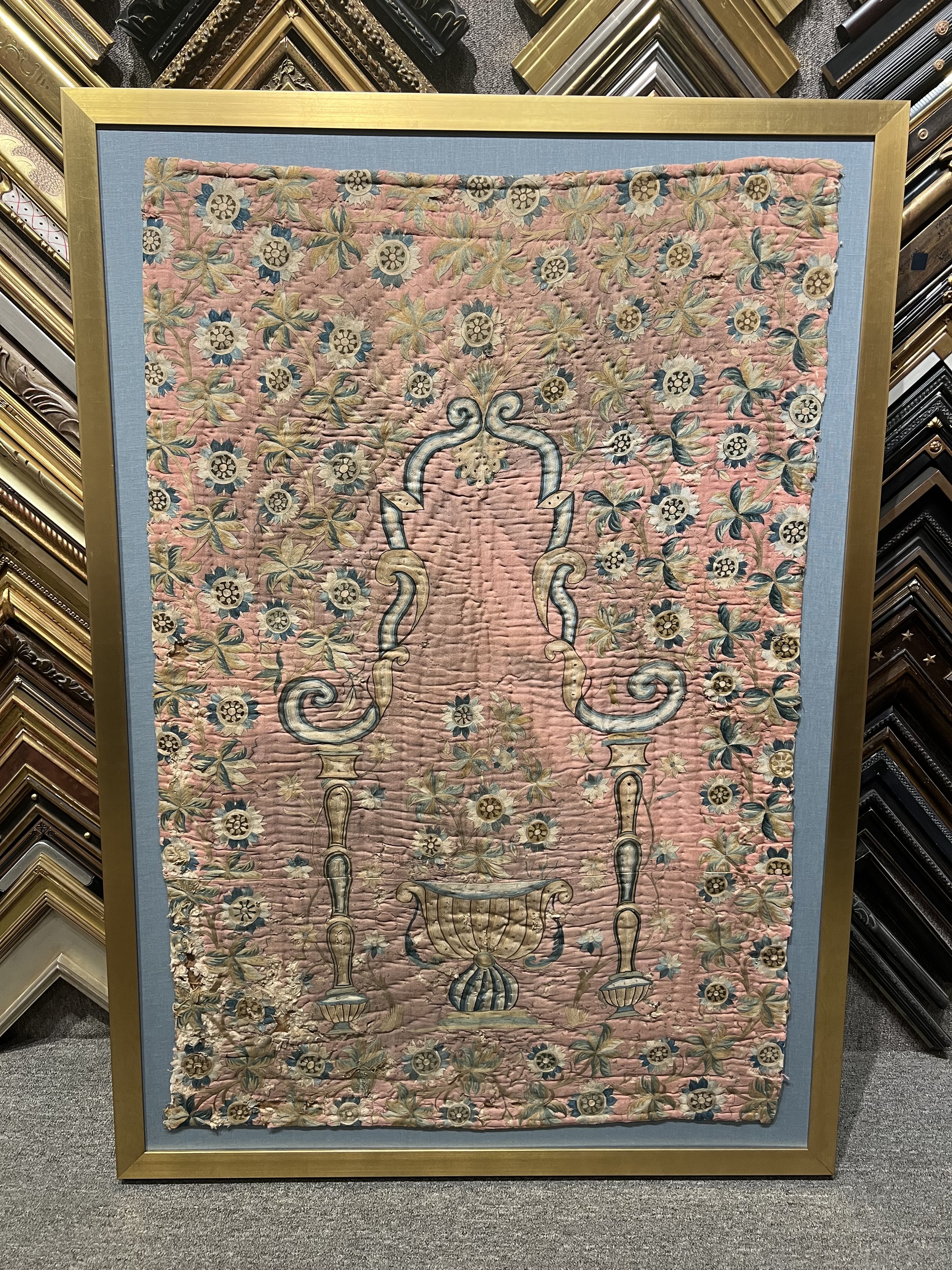 A vintage tufted tapestry with floral and urn motif in shades of pink, blue, and beige, framed in gold, set on a display wall surrounded by picture frame samples.