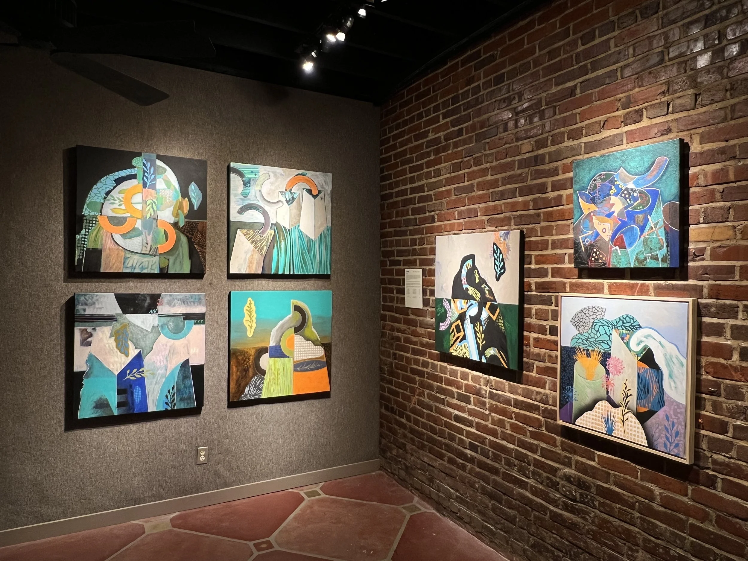 Art gallery with six abstract paintings on a gray wall and a brick wall, illuminated by ceiling spotlights.