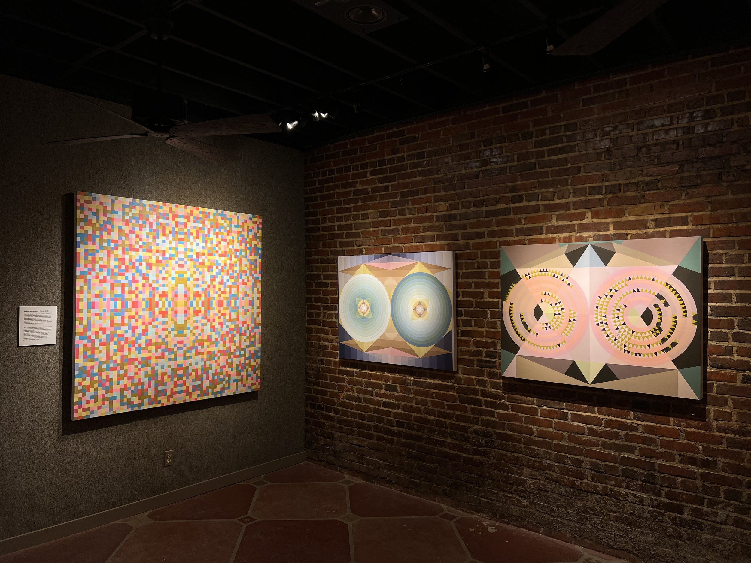 Gallery wall featuring abstract geometric artworks in a gallery with brick wall and spotlighting.