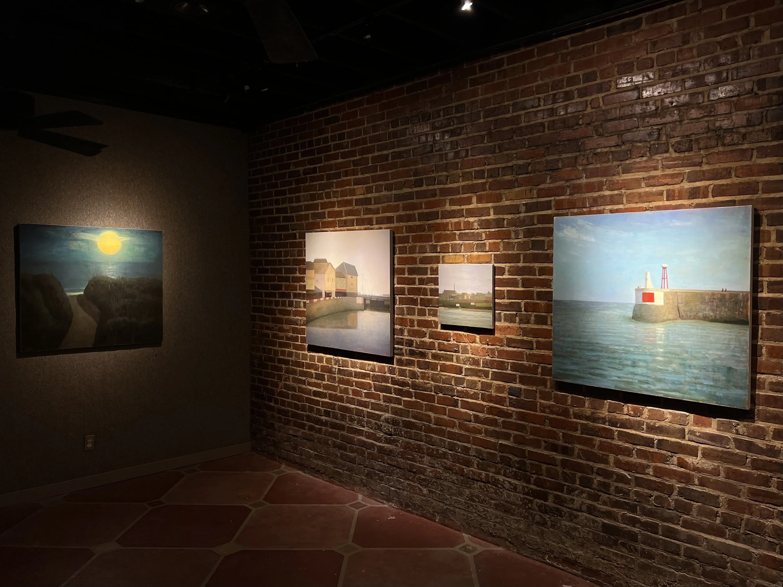Art gallery with paintings of seaside scenes on brick wall, dim lighting.