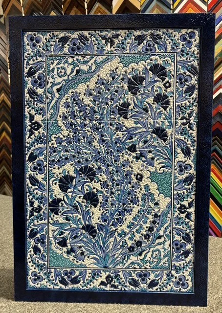 Decorative ceramic tile artwork featuring intricate blue and white floral patterns, stylized leaves, and small flowers surrounded by a dark border.