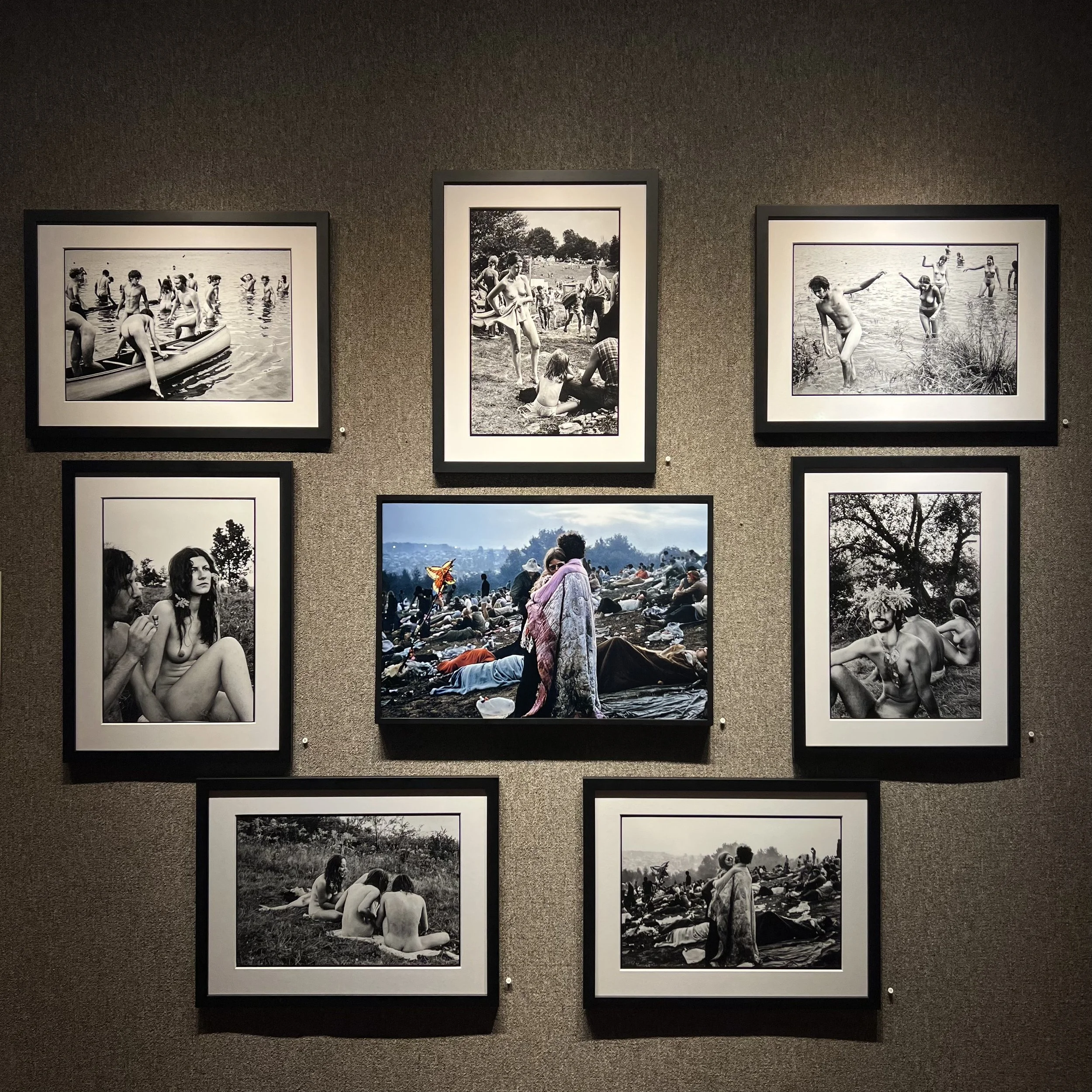 Black and white photographs displayed on a gallery wall depicting groups of people at the beach and outdoors, some in the water, others sitting and socializing in natural settings.