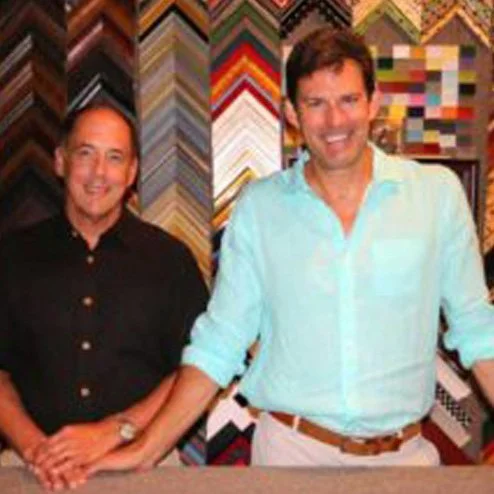Two men standing in front of a display of colorful picture framing samples, holding hands and smiling.