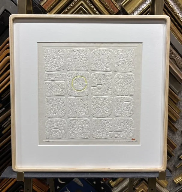 A framed white artwork displaying 16 embossed tiles with abstract patterns, including a yellow circle in the center, inside a white mat and wooden frame.