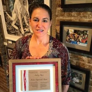 Woman holding a framed certificate or award indoors, with artwork on the wall behind her.