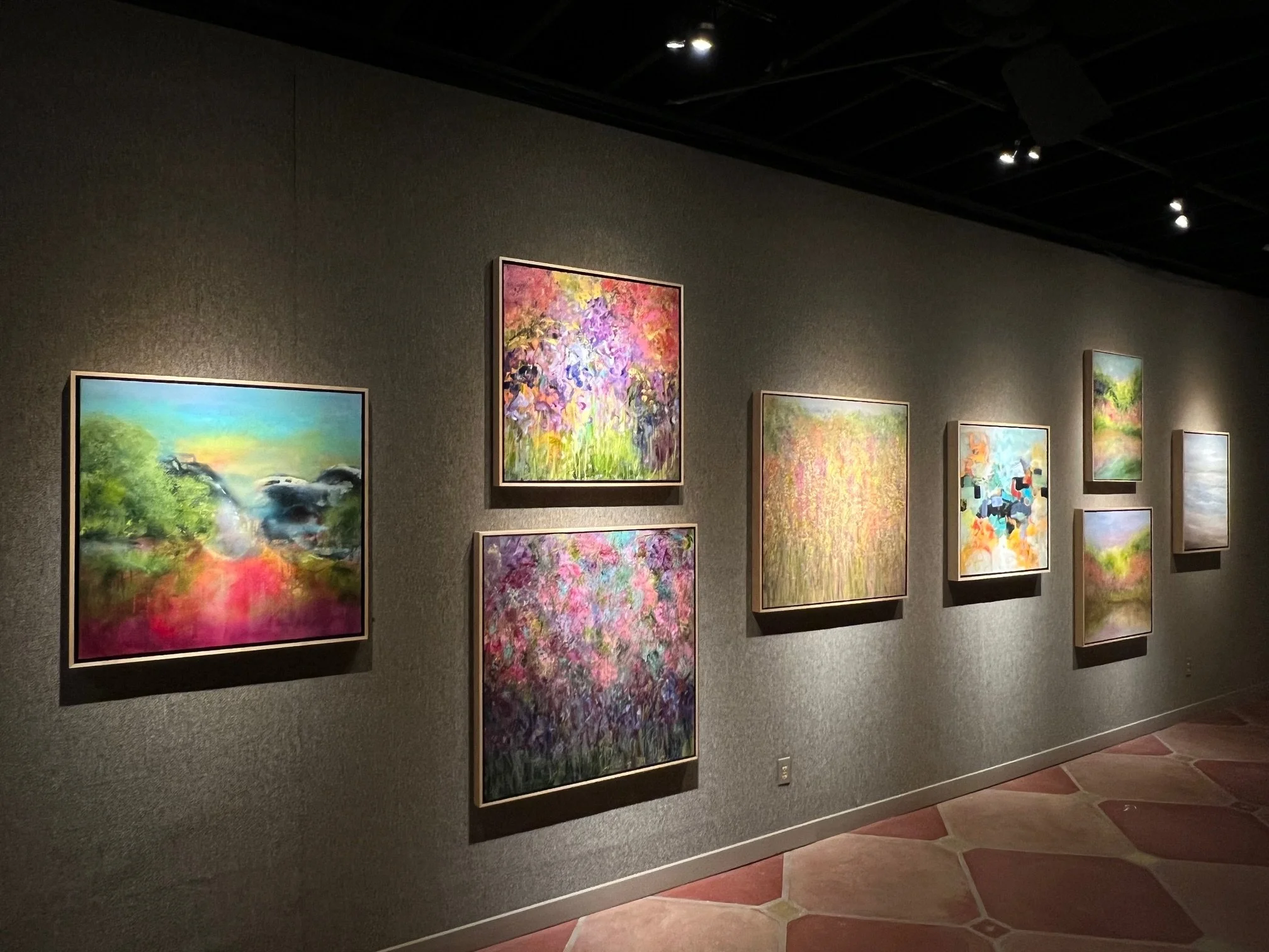 Art gallery wall displaying several colorful abstract paintings with a gray textured background and track lighting.