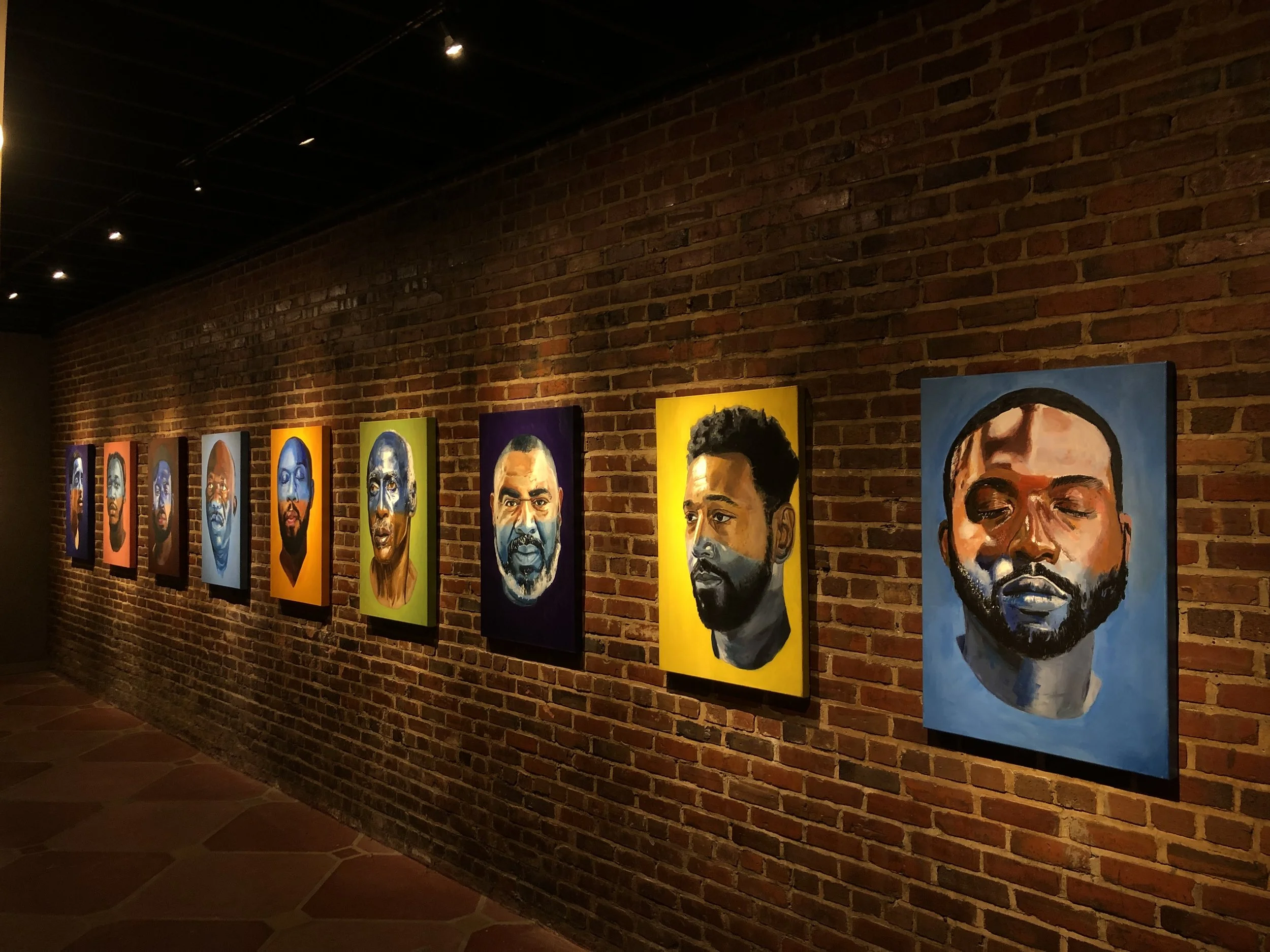 Gallery of colorful portrait paintings of diverse individuals, displayed on a brick wall in an art gallery with low lighting.