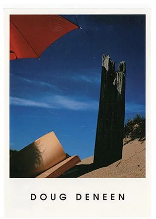 Photograph of a beach scene with a red umbrella, a wooden bench, a tall weathered wooden tower, and sand dunes against a night sky.