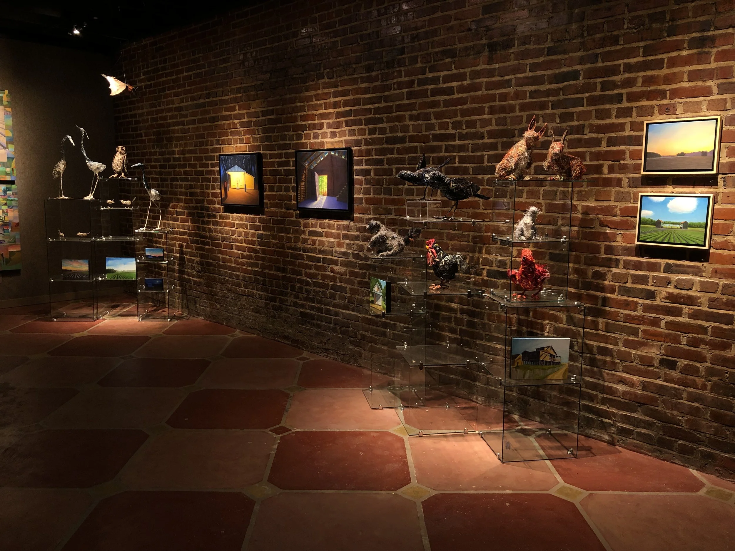 Art gallery display with sculptures of birds and animals on clear stands, along with framed landscape photographs on a brick wall.