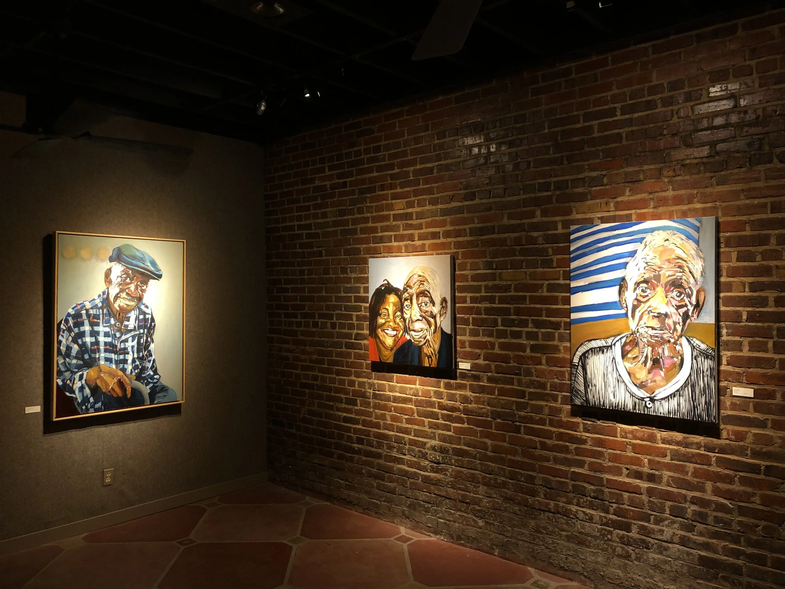 Art gallery with portrait paintings of elderly people on a brick wall.