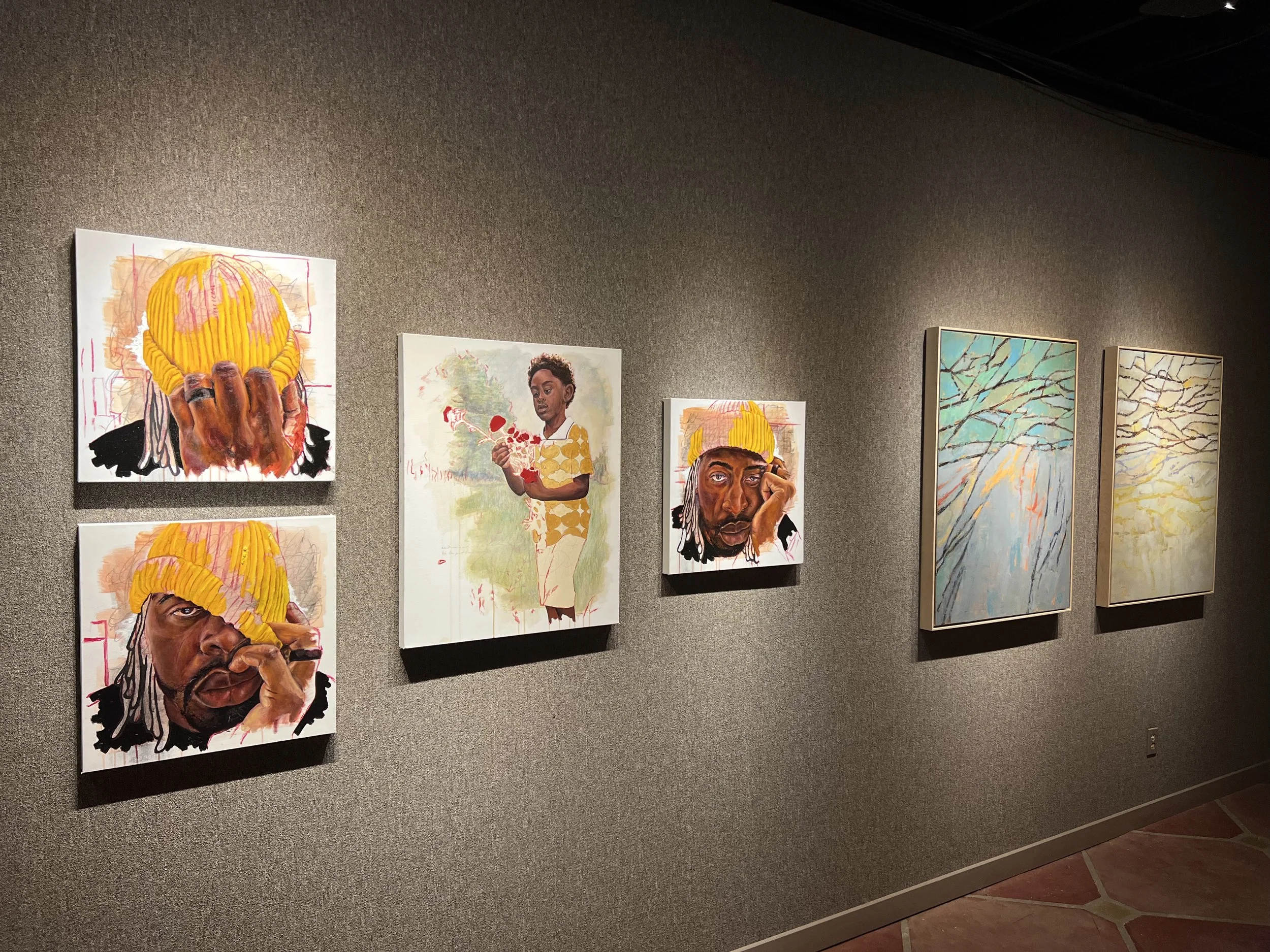 Art gallery wall displaying multiple paintings, including portraits of an African American woman and a man with dreadlocks, with landscapes featuring tree branches.