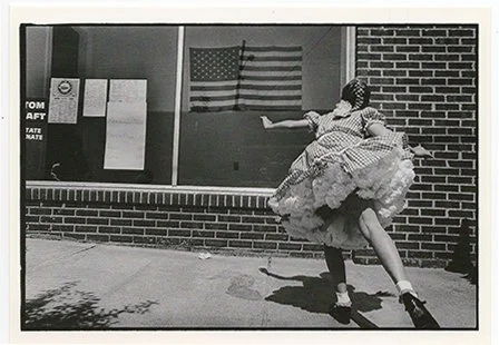 A person dressed in a frilly dress and hat, dancing or posing outdoors in front of a brick building with an American flag and bulletin board.