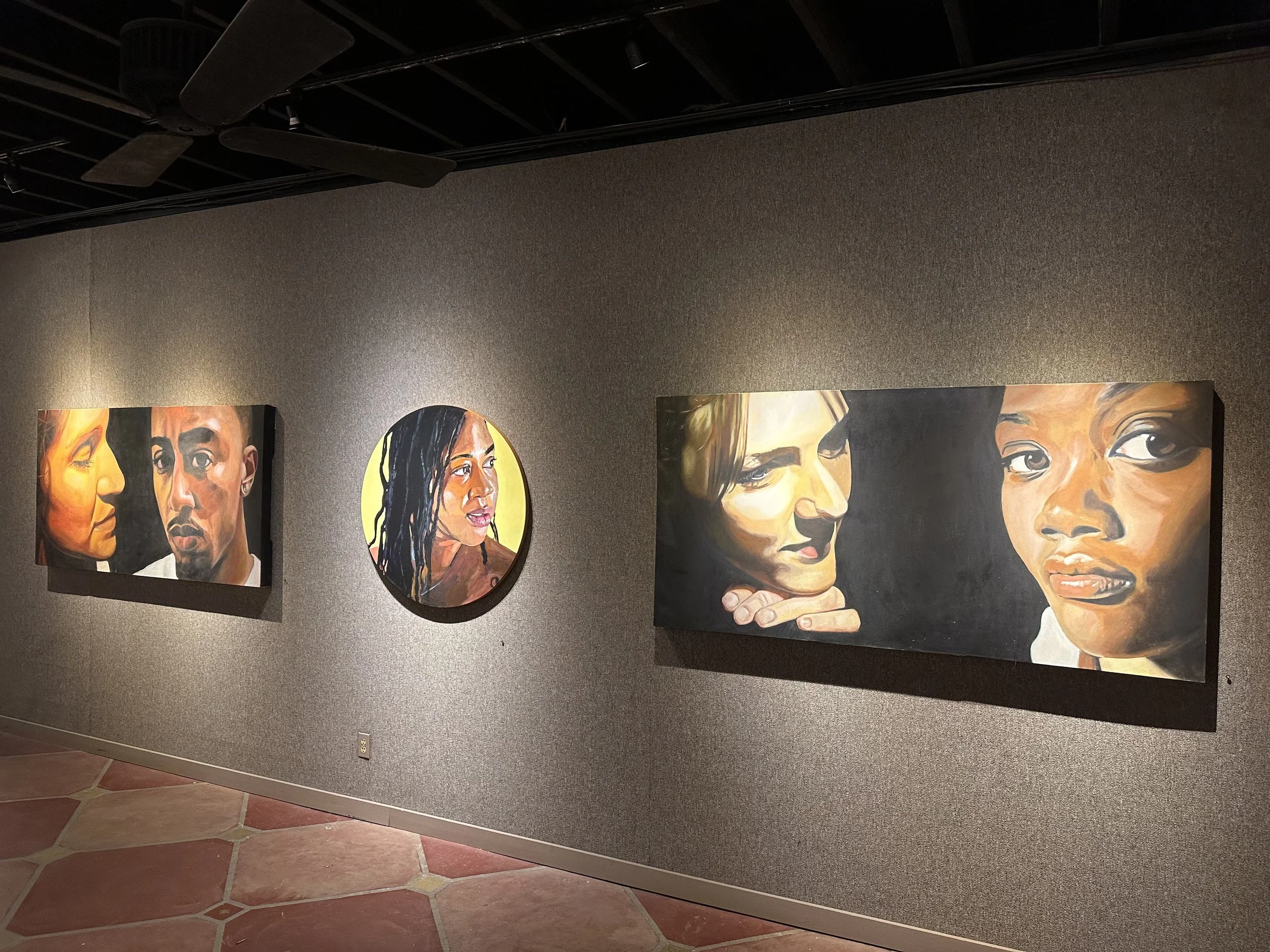Gallery wall displaying five colorful portrait paintings of diverse women with different expressions. The background wall is dark gray and the artworks are mounted in different shapes and sizes.