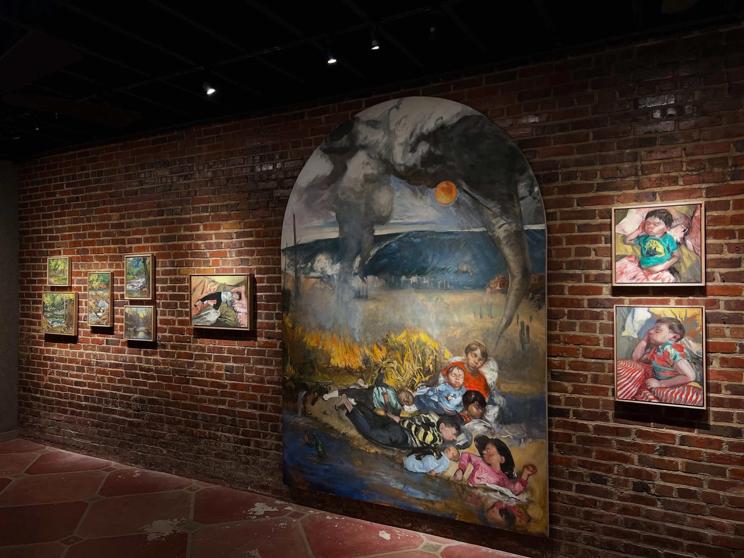 An art gallery with dark ceiling and brick wall, exhibiting paintings including a large mural of an elephant with a sunset background, and smaller portraits of children.