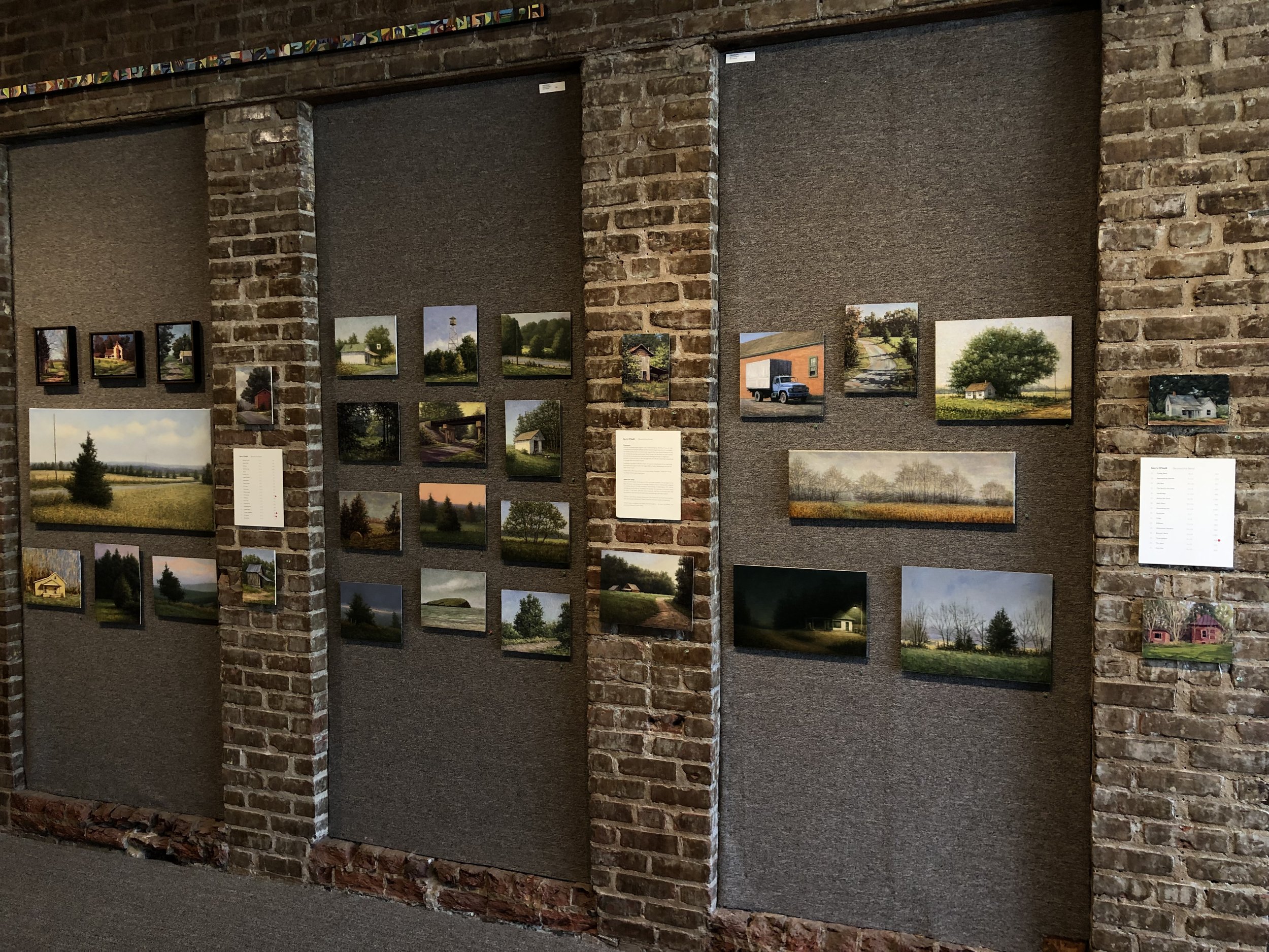 Gallery display of landscape photographs on a textured wall with brick columns, showing fields, trees, houses, and rural scenes.