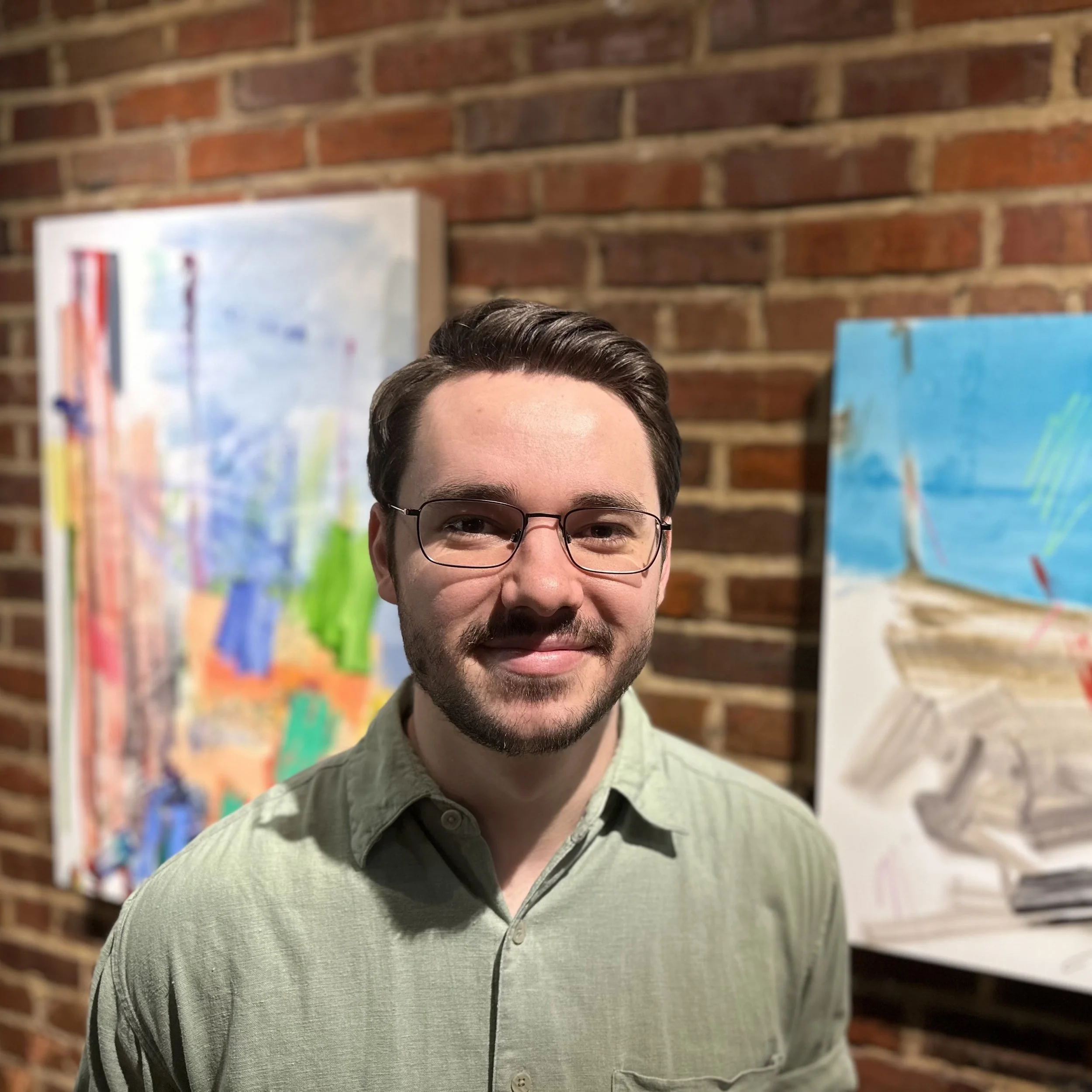 Man with glasses and short dark hair, smiling, wearing a light green shirt, standing in front of a brick wall with colorful abstract paintings.