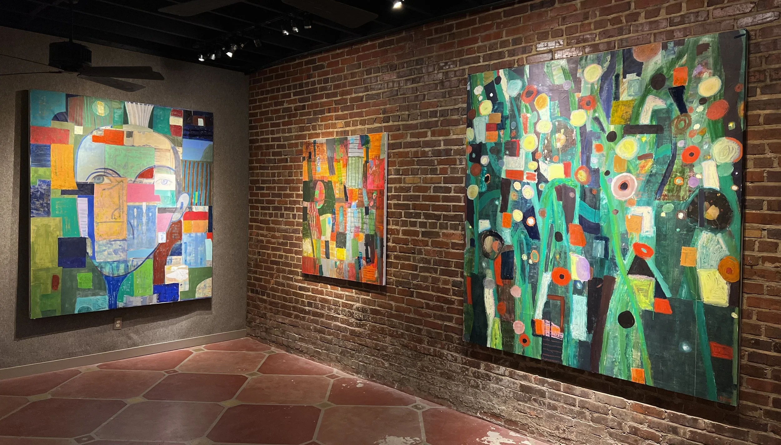 Three abstract colorful paintings on a brick wall, displayed in an art gallery with a tiled floor and ceiling lights.