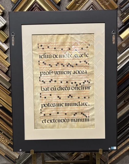 Framed ancient manuscript of a musical score with Latin text, displayed in a gallery among other frames.