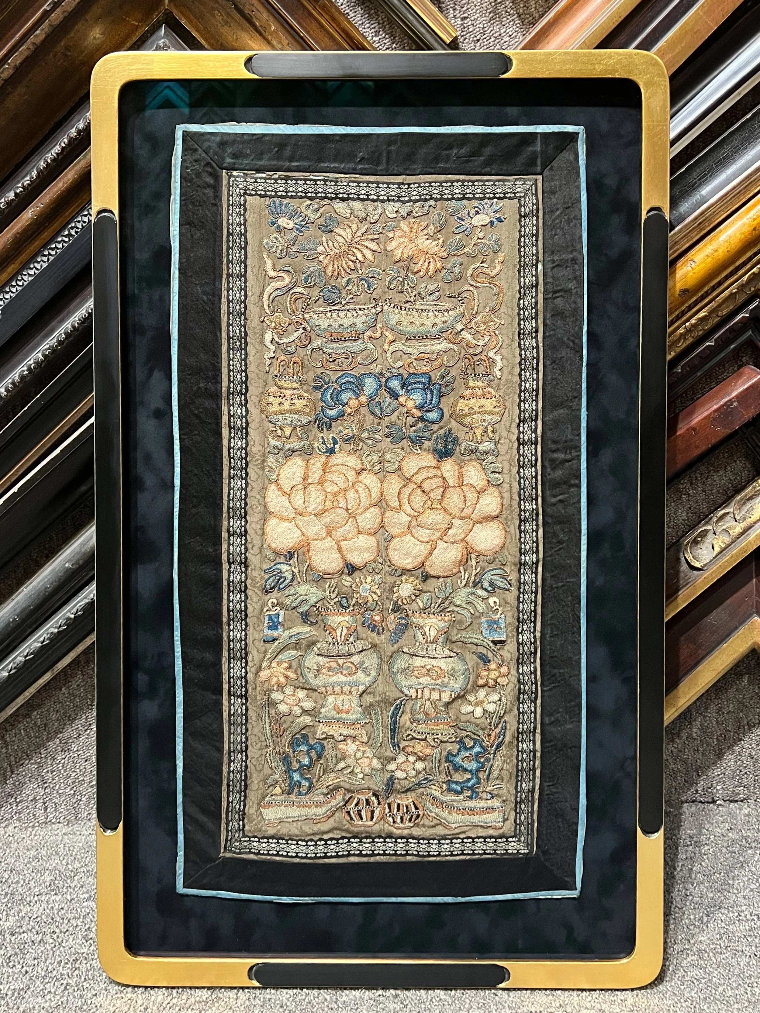 An ornate, embroidered Asian-style rug with floral and dragon motifs, displayed on a black and yellow cart in a store with various other framed rugs stacked around.