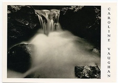 A black and white photo of a waterfall flowing between rocks in a river or stream, with the name Caroline Vaughan written vertically to the right.